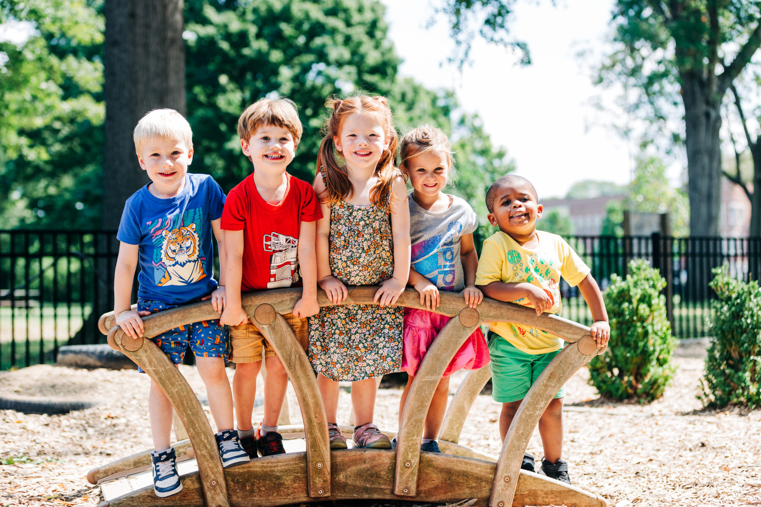 Five Child Development Center children smiling outside on a wooden bridge on their natural playground